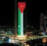 Nighttime city skyline with a tall, illuminated green-and-white tower topped with a red emblem, surrounded by other buildings.