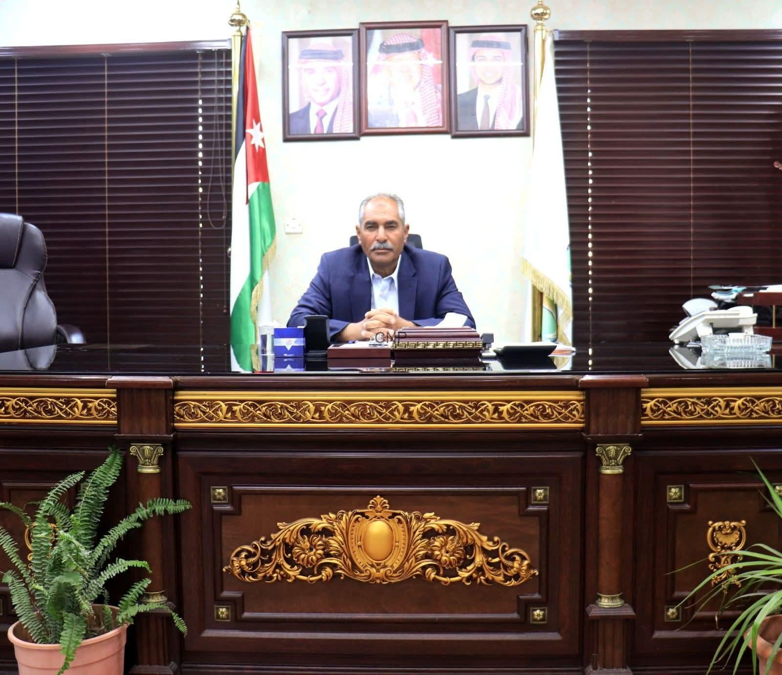 Man in a blue suit seated at a large ornate wooden desk in a formal office, with a Jordan flag and framed portraits on the wall behind him.