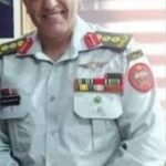Smiling man in a light gray uniform with ribbons and patches, posing indoors.