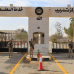 Border checkpoint entrance with three arches, armed soldiers standing guard, a small guardhouse between arches, yellow lane markings, and an orange traffic cone; Arabic signs and crown logos on the facade.