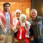 Four people posing together indoors; two men on the ends wear casual clothes and red-and-white keffiyehs, while two women in white hijabs stand between them against an orange patterned wall.