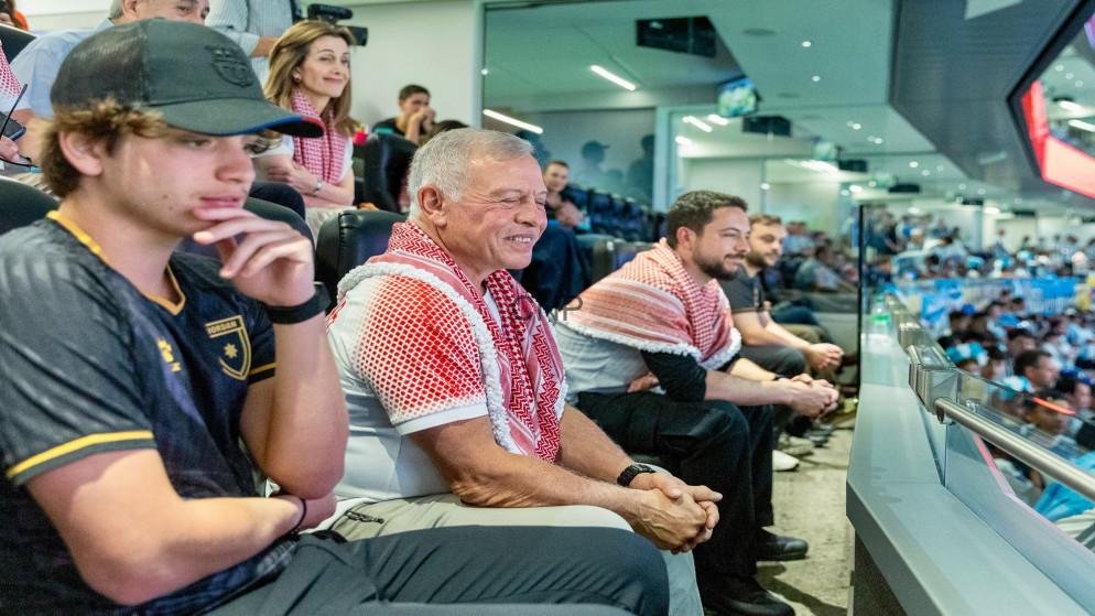 Older man smiling in a red-and-white keffiyeh scarf seated among spectators in a VIP-style arena box.