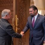 Two men in suits shake hands at a carved wooden door in a formal lobby setting.