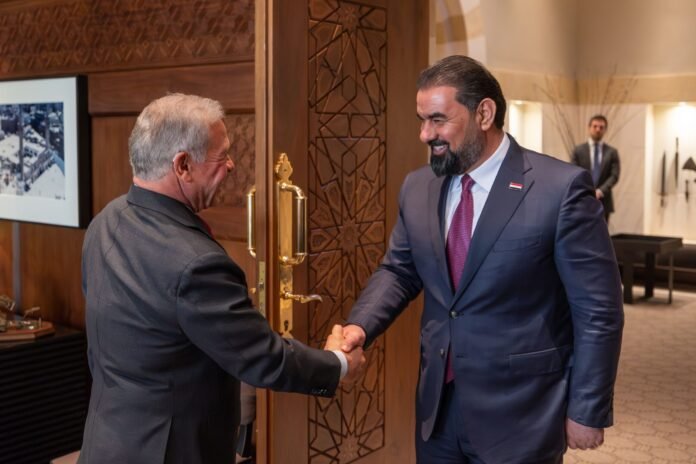 Two men in suits shake hands at a carved wooden door in a formal lobby setting.