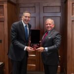 Two men in suits exchange a red velvet medal box, smiling, in a wood-paneled room.
