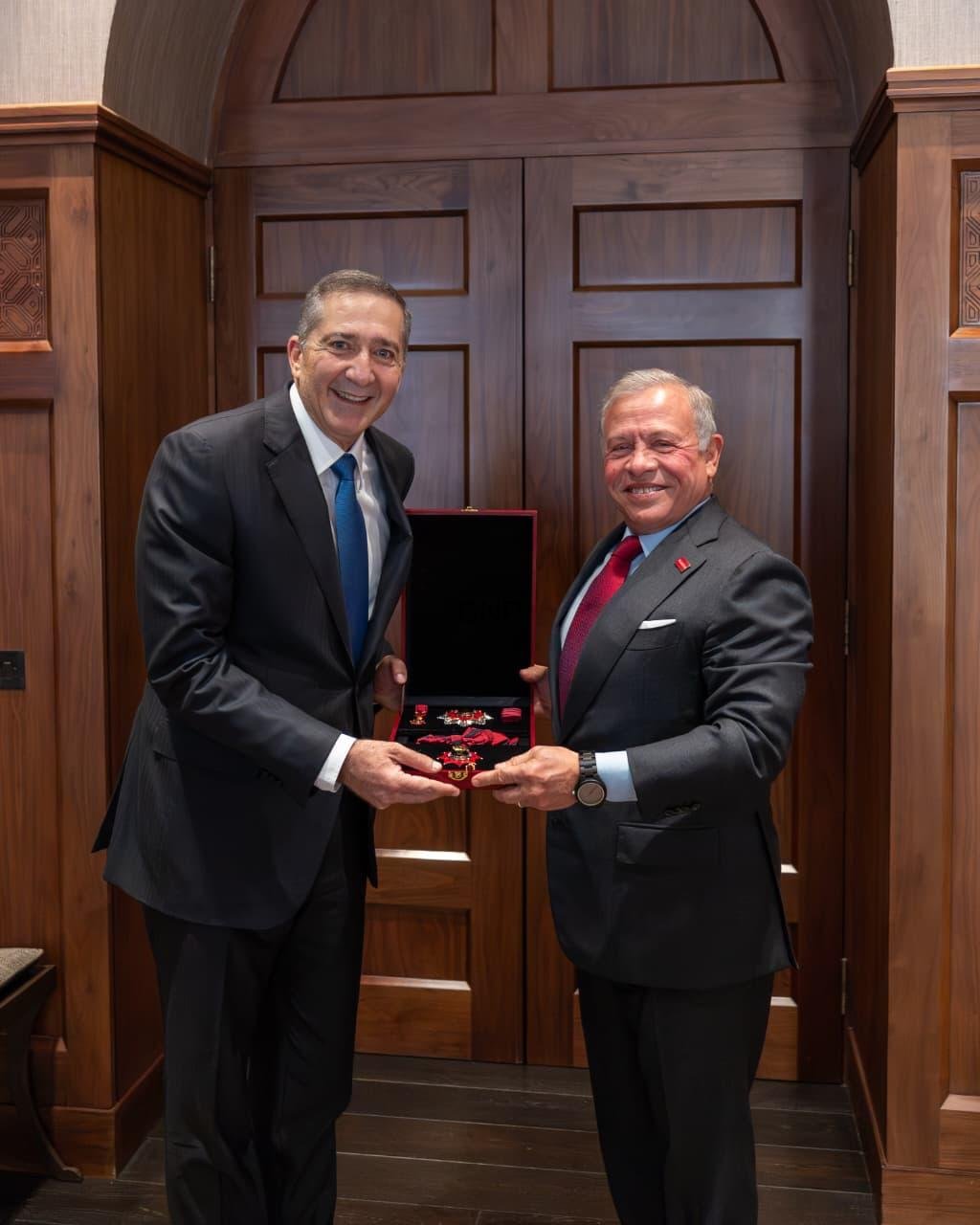 Two men in suits exchange a red velvet medal box, smiling, in a wood-paneled room.