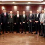 Twelve men in suits standing in a formal meeting room with wooden paneling and flags in the background.
