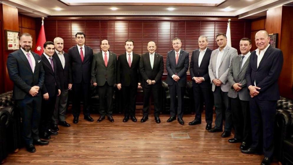 Twelve men in suits standing in a formal meeting room with wooden paneling and flags in the background.
