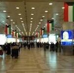 Busy airport terminal concourse with travelers walking under hanging Italian flags and digital information screens on the walls and screens above.