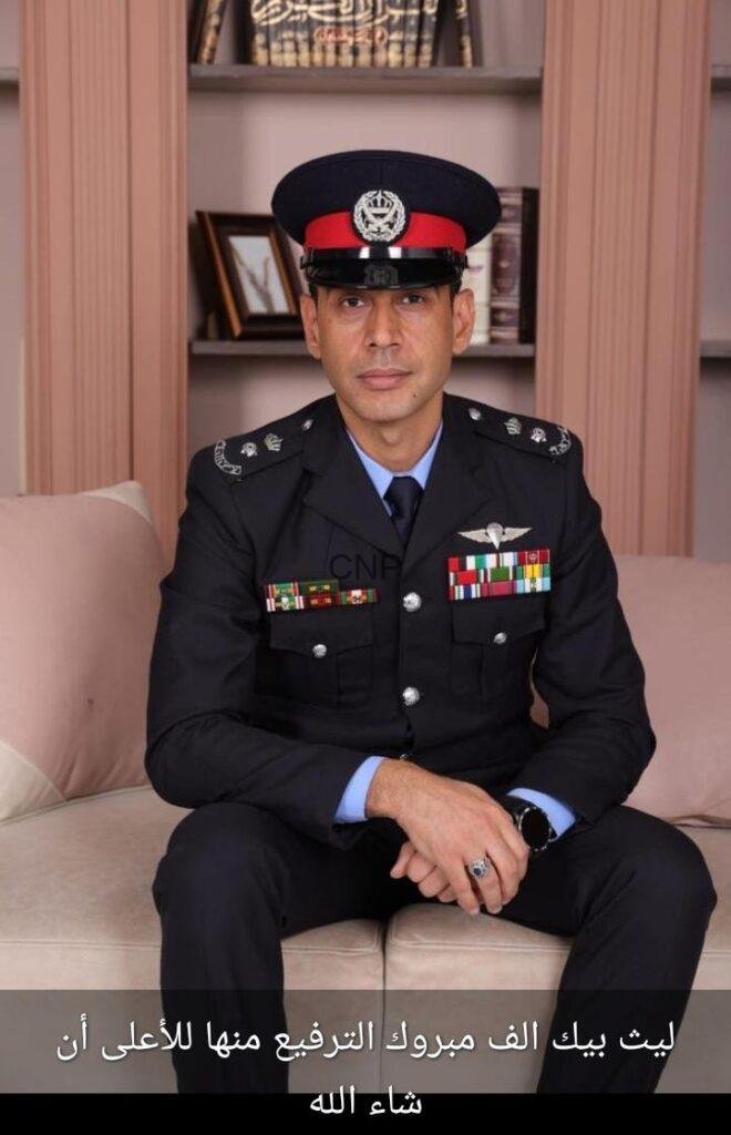laits - Center News Network Portrait of a male military officer in full dress uniform with medals, seated on a beige sofa, wearing a peaked cap with a red band, in a room with shelves behind him; hands folded in his lap and a badge on the chest are visible.