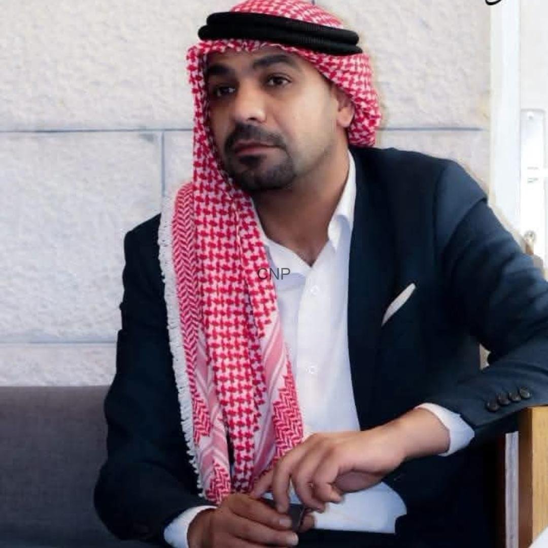 mohan - Center News Network Man seated on a sofa wearing a red-and-white keffiyeh and a dark blazer, looking toward the camera in a formal pose.