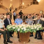 Group of officials and military officers seated at a formal ceremony around a flower-adorned table in a grand hall.