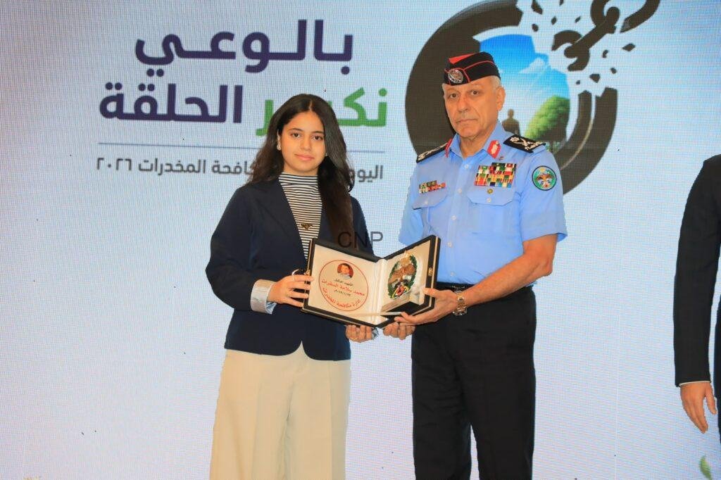 mok2 - Rakeen News Young woman in a navy blazer receives a certificate from a uniformed officer at an awards ceremony on stage, with Arabic text backdrop.