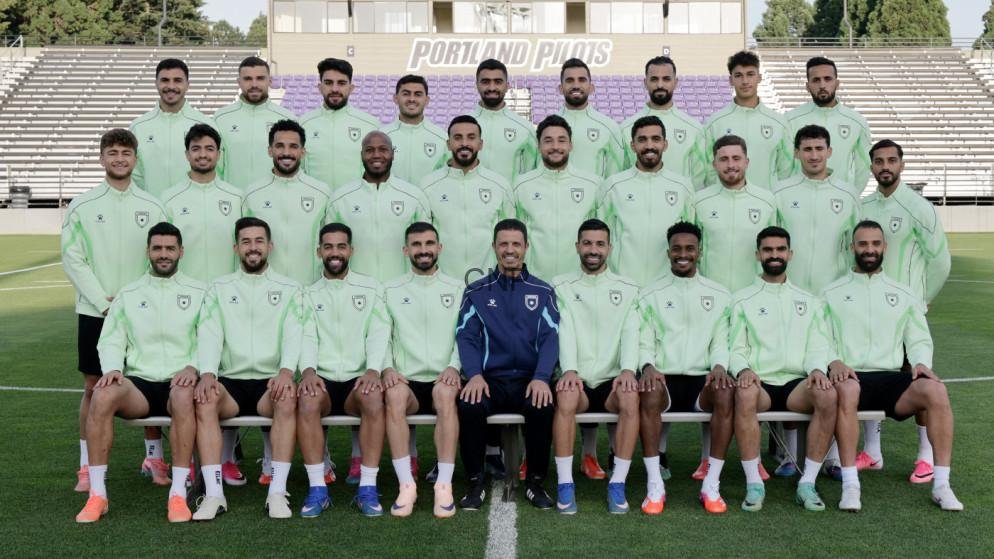 monnn - Center News Network Group photo of a soccer team posing on a stadium field in matching light green training jackets; a coach in a dark tracksuit sits center front, bleachers and a 'Portland' sign are in the background.
