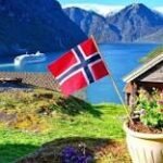 Norwegian flag by a lakeside cabin, with flowers, a boat on the fjord and snow-capped mountains in the background.
