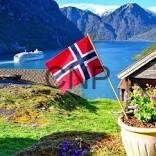 nar - Center News Network Norwegian flag by a lakeside cabin, with flowers, a boat on the fjord and snow-capped mountains in the background.
