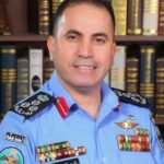 Portrait of a male officer in a light blue uniform with multiple ribbons and insignia, standing in front of a bookshelf full of legal tomes for an official portrait