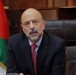 Bald, bearded man in a dark suit speaking from a desk in a formal setting, with a flag in the background.