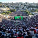 Thousands of spectators fill a sunlit outdoor amphitheater, watching a large televised football match on a central screen.