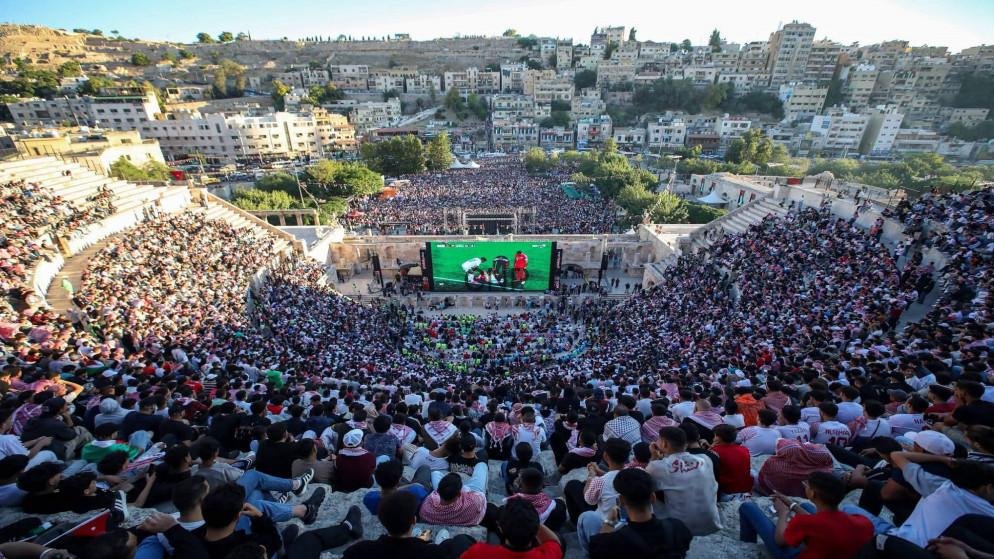 Thousands of spectators fill a sunlit outdoor amphitheater, watching a large televised football match on a central screen.