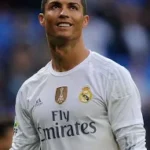 Male soccer player in a light gray Real Madrid jersey with Fly Emirates on the chest, standing on a field.