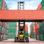 Forklift lifting a red shipping container between stacked multicolored containers in a cargo yard.