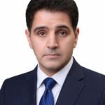 Professional head-and-shoulders portrait of a man in a navy suit and blue tie with neutral expression, white background.