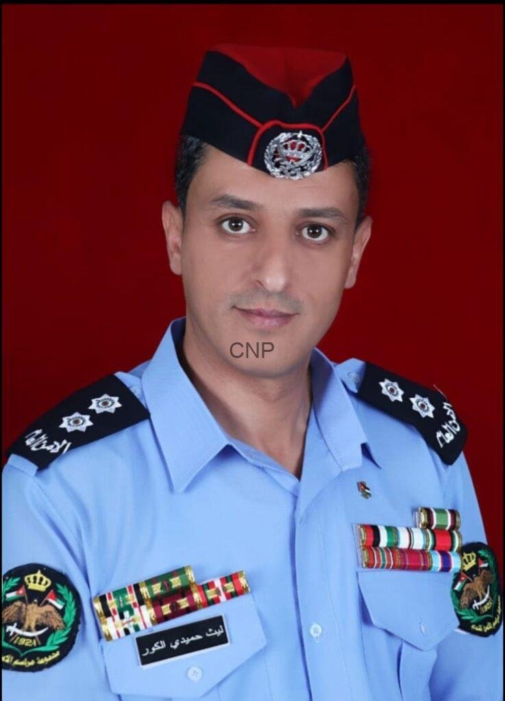 slait - Center News Network Portrait of a man in a light blue ceremonial uniform with medals and shoulder insignia, wearing a black cap with red trim against a red background.