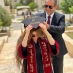 Man in a suit and sunglasses places a graduation cap on a woman in a black dress and maroon stole outdoors on a campus-like walkway, cap with gold Arabic text visible.