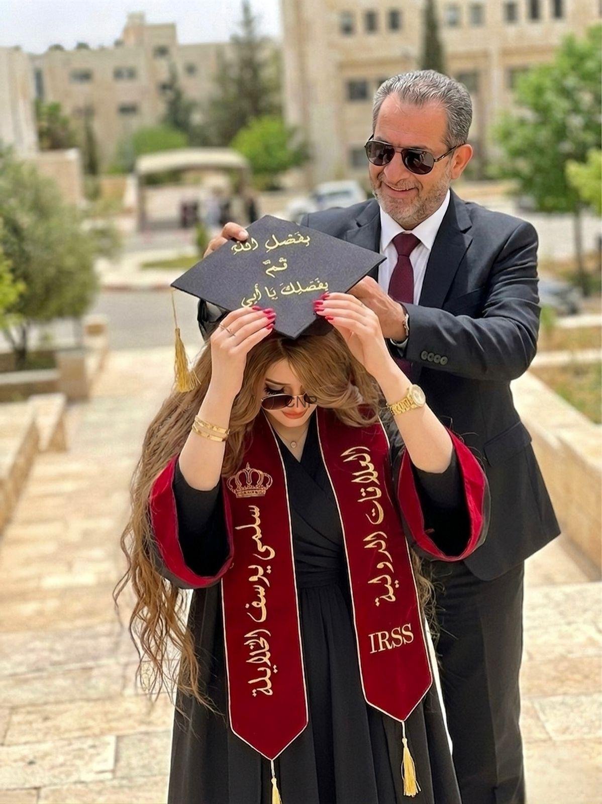 sobh - Center News Network Man in a suit and sunglasses places a graduation cap on a woman in a black dress and maroon stole outdoors on a campus-like walkway, cap with gold Arabic text visible.
