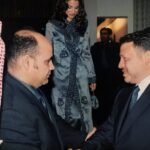Two men in dark suits shake hands at a formal event, with a woman in a blue embellished outfit visible in the background.