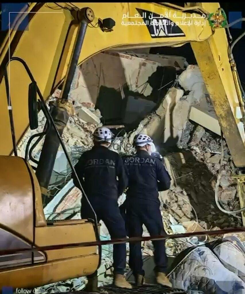 Two rescue workers in dark uniforms and white helmets stand amid rubble from a collapsed building, next to a yellow excavator arm during a nighttime search.