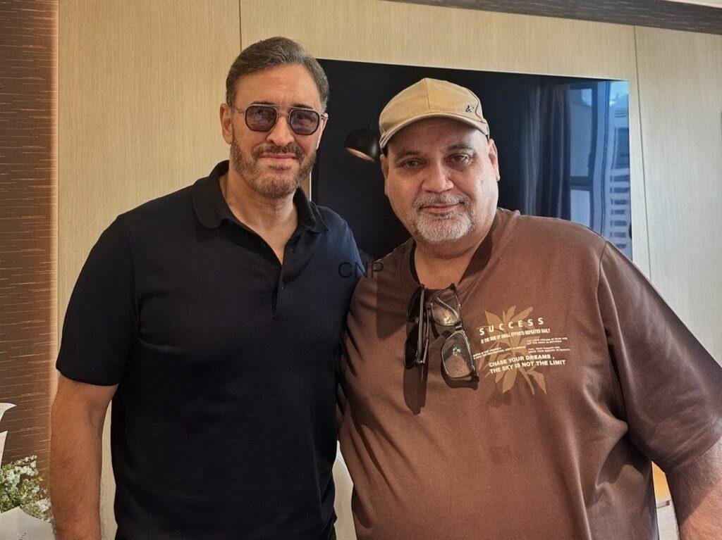 ssaaddf - Center News Network Two men pose indoors for a photo; one in a black polo with sunglasses, the other in a brown sweatshirt and tan cap, both smiling.
