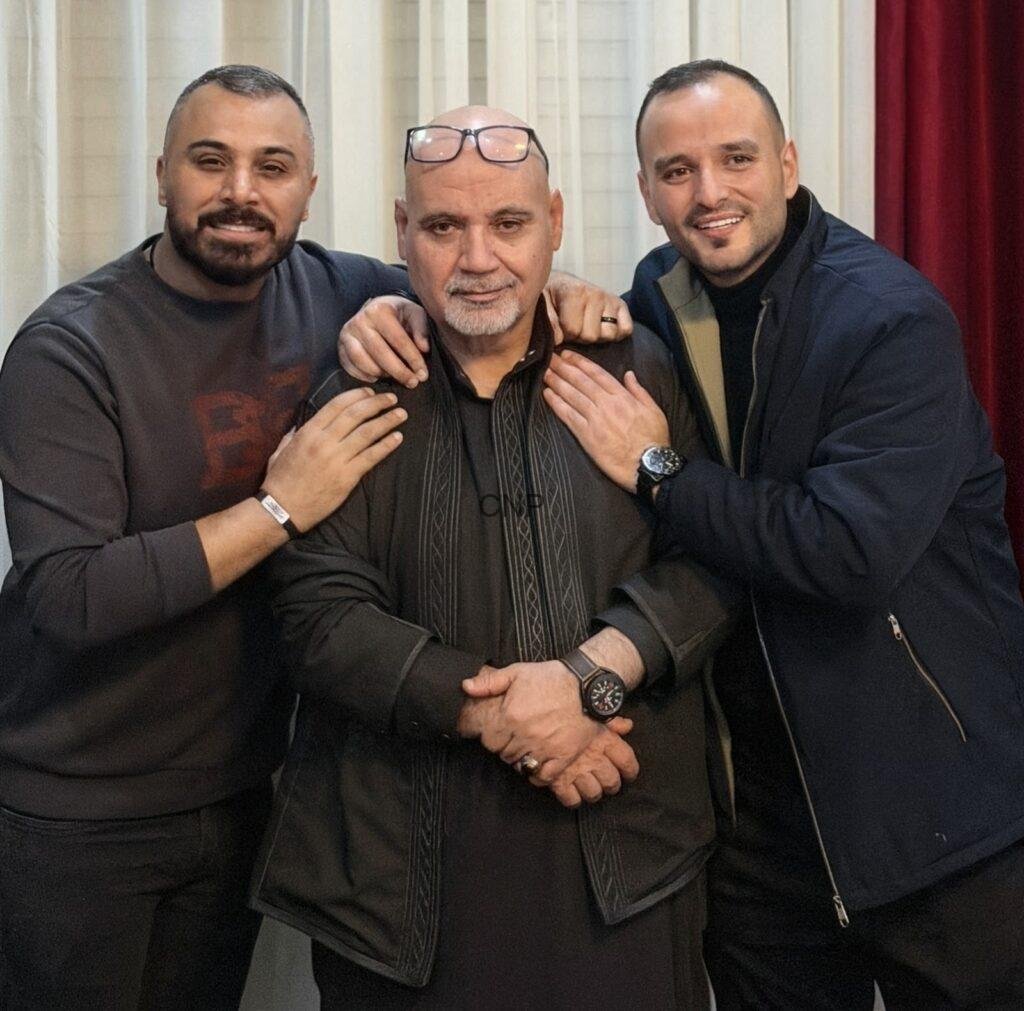 sss17 - Center News Network Three men posing closely for a group photo, with the middle older man wearing a black traditional outfit and glasses on his head, flanked by two smiling friends with their arms around him.