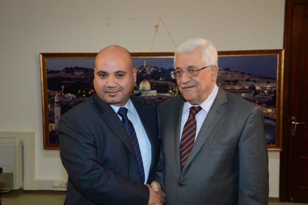 sss55 - Center News Network Two men in suits shake hands in front of a framed cityscape photo in an office setting.