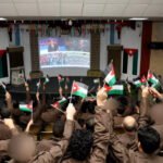 Audience in brown uniforms waving Palestinian flags while watching a broadcast on a large screen in a decorated hall with flags on the walls and ceiling.