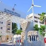 Entrance to a campus or corporate complex featuring a curved stone gate with a large sign, a wind turbine in the background, and a beige multi-story building under a clear blue sky.