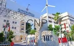 Entrance to a campus or corporate complex featuring a curved stone gate with a large sign, a wind turbine in the background, and a beige multi-story building under a clear blue sky.