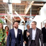 Group of professionals walking and chatting in a bright, open office with exposed ceilings and ductwork. A man in a blue suit smiles as he walks beside a gray-suited colleague who gestures while speaking.