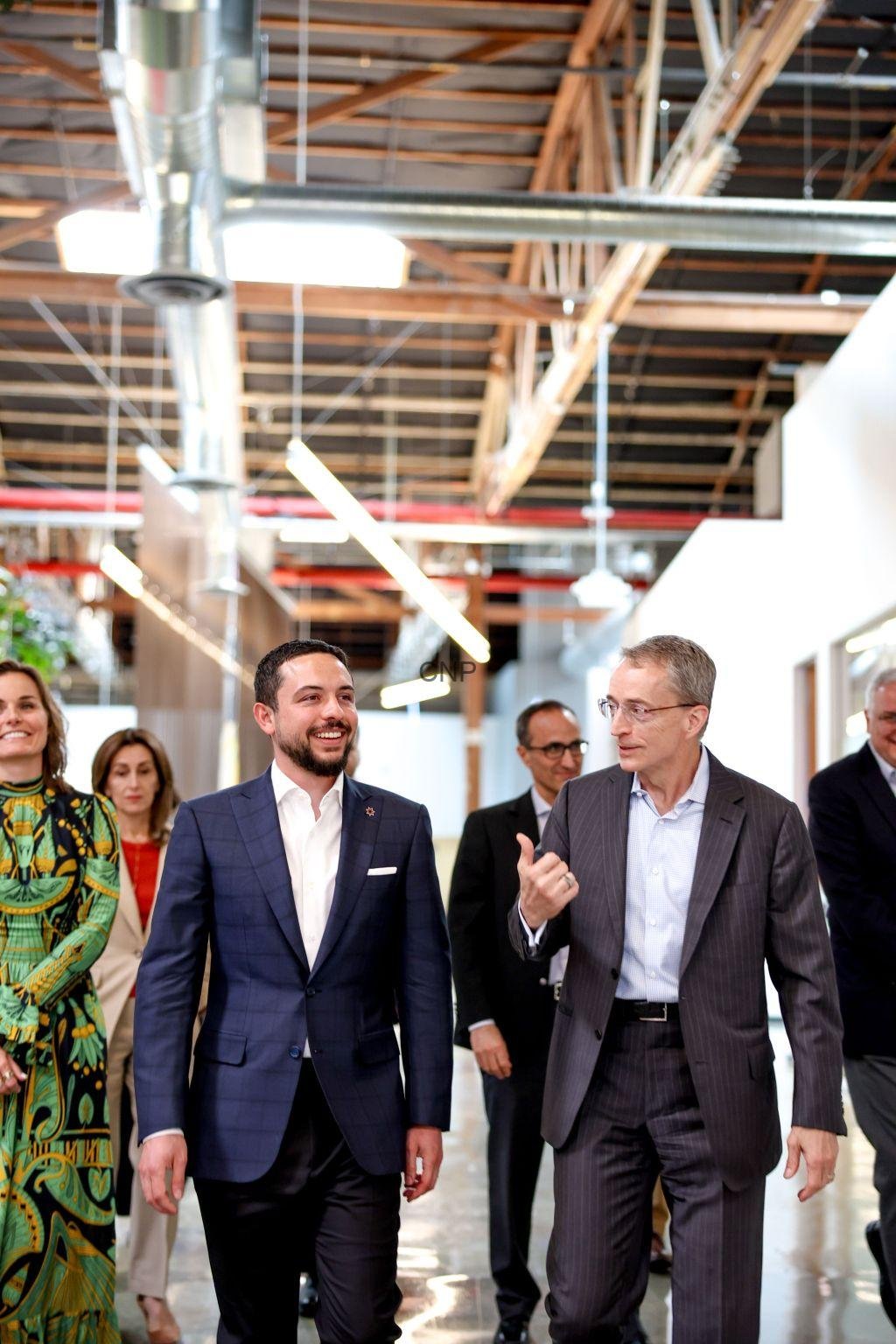 waly - Center News Network Group of professionals walking and chatting in a bright, open office with exposed ceilings and ductwork. A man in a blue suit smiles as he walks beside a gray-suited colleague who gestures while speaking.