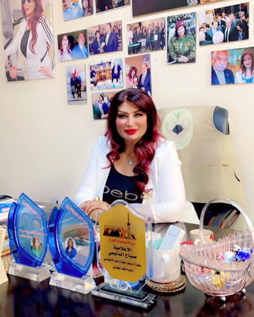 Smiling woman in a white blazer sits at a desk displaying multiple blue trophy plaques; wall behind her is covered with photo collages and awards photos.