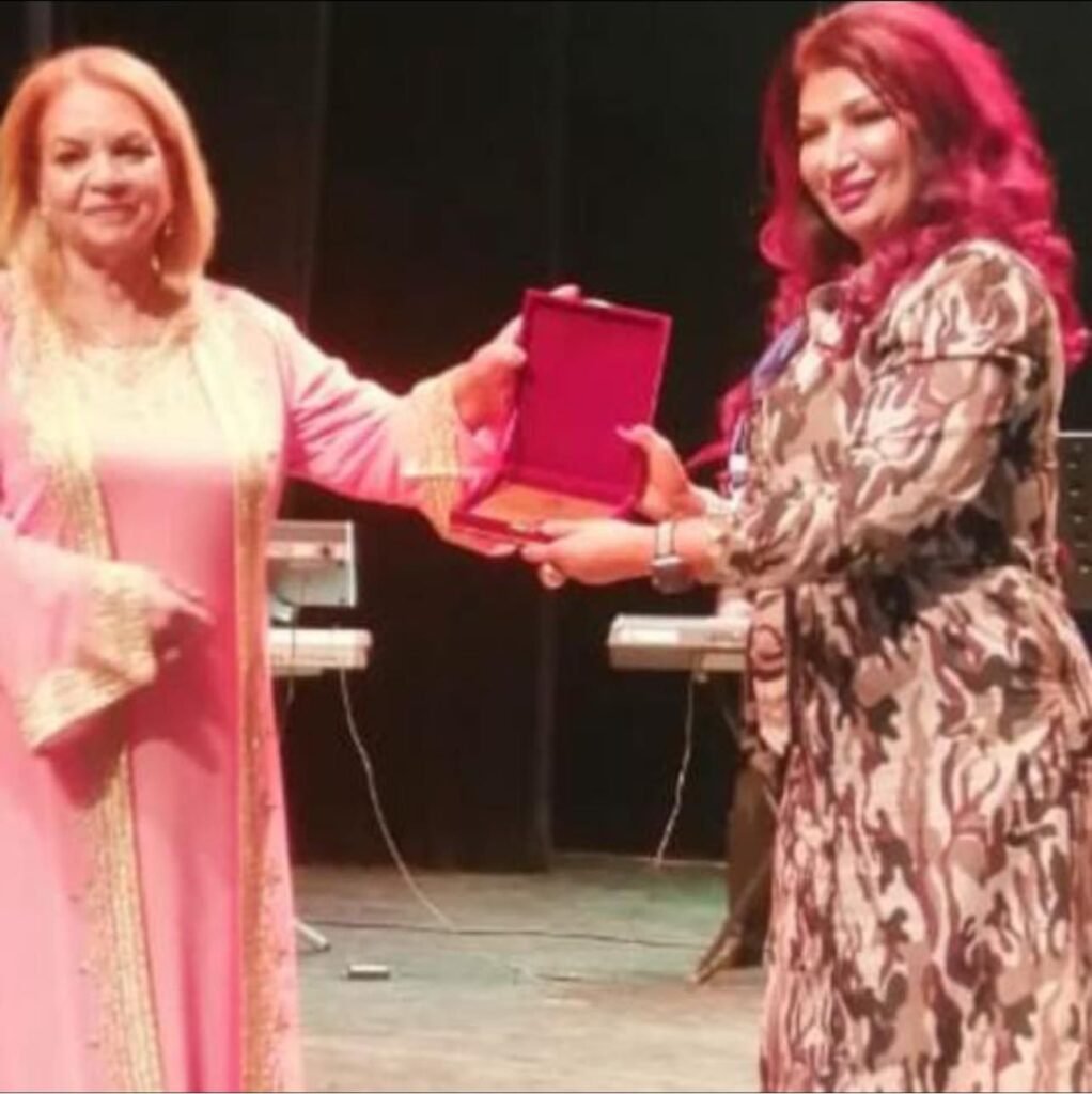 Two women on a stage exchange an open red award box during a ceremony, both smiling.