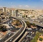 Aerial view of a complex multi-level highway interchange weaving through a dense urban area with beige buildings under a blue sky.