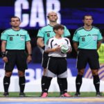 Six referees in teal uniforms stand in a line on a soccer field, with a young boy holding a ball in front of them.