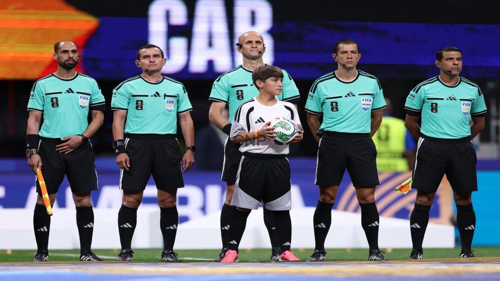 Six referees in teal uniforms stand in a line on a soccer field, with a young boy holding a ball in front of them.
