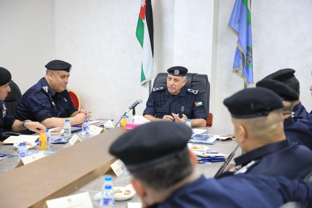 Senior police officer in uniform seated at head of conference table, leading a formal meeting with colleagues, flags behind him.
