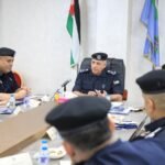 Senior police officer in uniform seated at head of conference table, leading a formal meeting with colleagues, flags behind him.