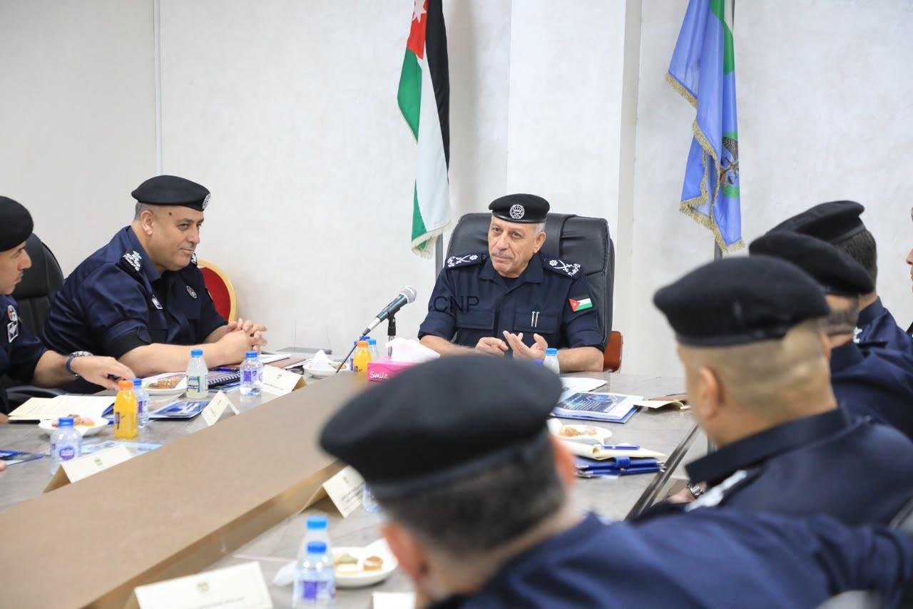Senior police officer in uniform seated at head of conference table, leading a formal meeting with colleagues, flags behind him.