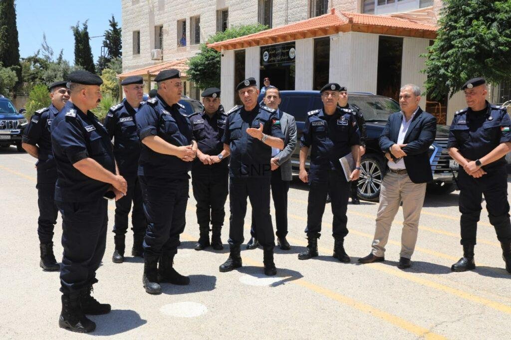 Group of uniformed police officers listening as a speaker talks outdoors in a parking lot outside a building, with cars nearby.