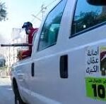 White emergency vehicle with Arabic text and 102, a firefighter stands on its rear step as smoke billows nearby.